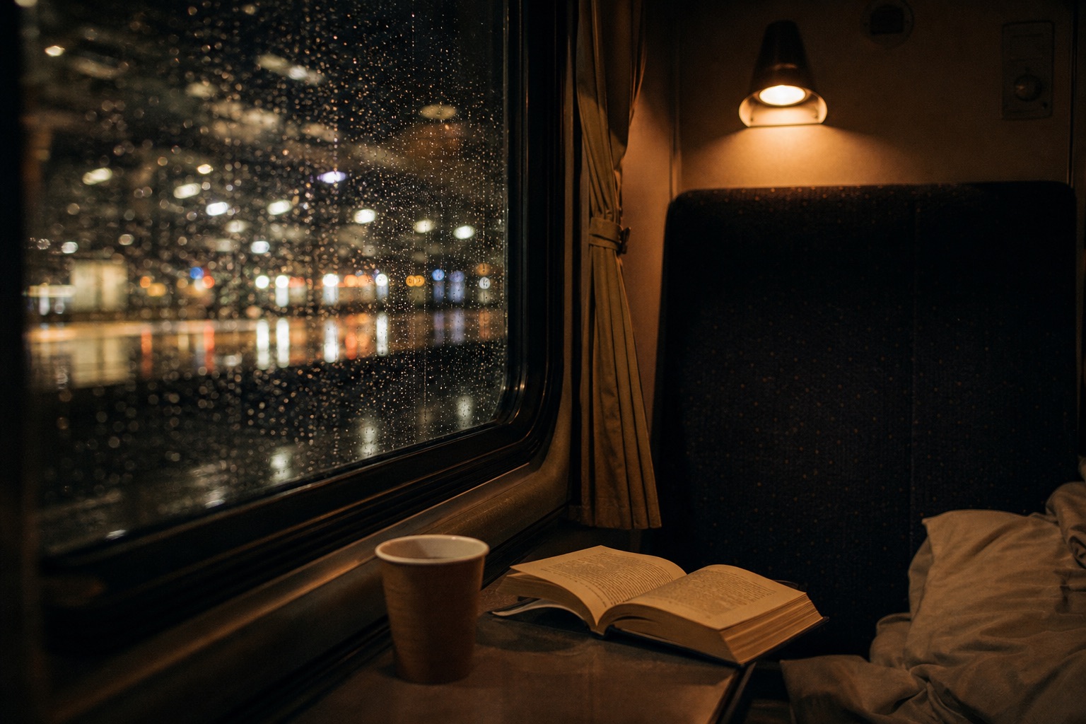An open book and paper cup beside a rain-covered night train window